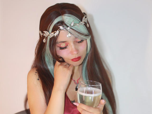 YukkyKuran with long brown and green hair, wearing a silver leaf headpiece, red lipstick, and a pink tank top, holding a glass of beer, looking down with a gentle smile.