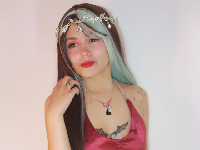 YukkyKuran with long brown and green hair, wearing a pink satin dress, silver headpiece, red lipstick, and necklace, has a bat tattoo on her chest, and a ring.