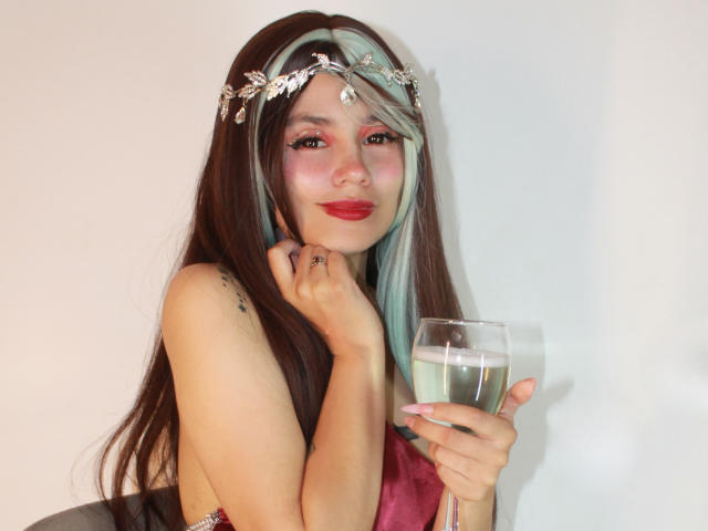 YukkyKuran with long brown and teal hair, wearing a jeweled headband, red lipstick, holding a glass of green drink, smiling, showing her tits and a tattoo on her shoulder.