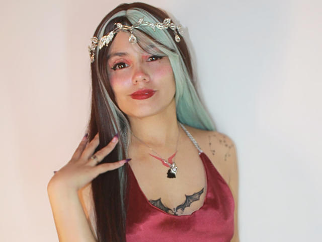 YukkyKuran with long brown and green hair, red lipstick, and a red satin dress. Wearing a floral headpiece, necklace, and showing tattoos on her chest. Hands up.