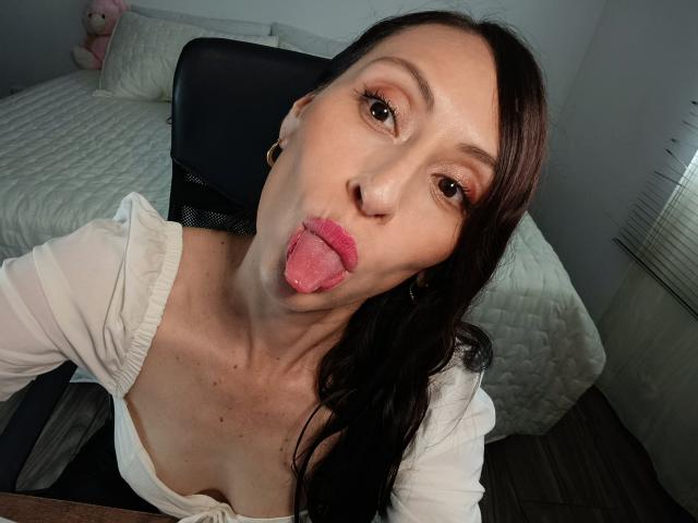 AliceRossi with pouty pink tongue, brown eyes, and long dark hair, wearing a white top, sitting in a black office chair, showing her cleavage and small earrings.