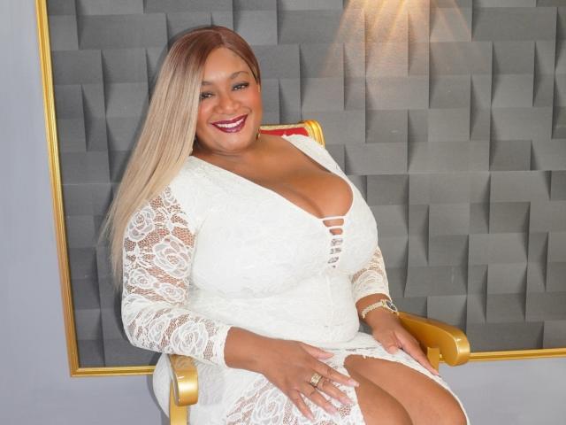 NahomiAnders with big tits, wearing a white lace dress, sitting on a golden chair, smiling with red lipstick, blonde hair, and a gold bracelet.