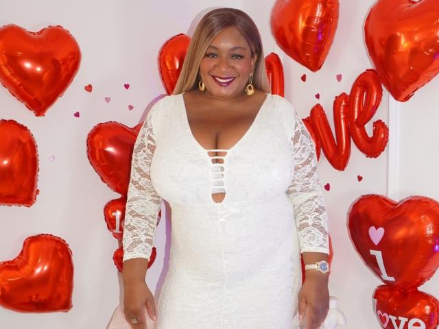 NahomiAnders with big boobs in a white lace dress, smiling, standing in front of red heart balloons. She's wearing gold earrings and a watch. Her hair is blonde.