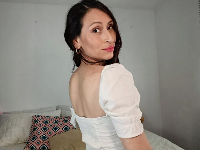 AliceRossi has medium-length black hair, light skin, and brown eyes. She's looking over her shoulder with a slight smile. She's wearing a white, off-shoulder, puffed-sleeve top. Her right ear has a gold hoop earring. She's sitting on a bed with a beige cover and a pink, geometric-patterned pillow. The room has plain, white walls. The image is well-lit with soft, natural light.