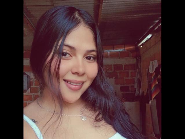 Sicaru has long black hair, brown eyes, and a cute smile with pink lips. She's wearing a white top and a necklace with a small pendant. Her tits are small.
