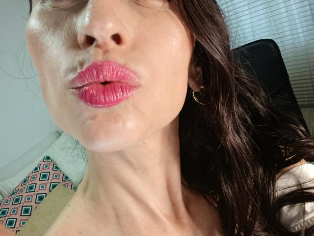 AliceRossi's face with glossy pink lips, slightly open, showing a hint of teeth. She has fair skin, dark wavy hair, and gold earrings. Her neck is visible.
