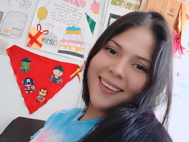 Sicaru, with long black hair, smiling, wearing a blue tie-dye shirt, has cute, small tits, and a cute face, standing near colorful, superhero-themed wall art.