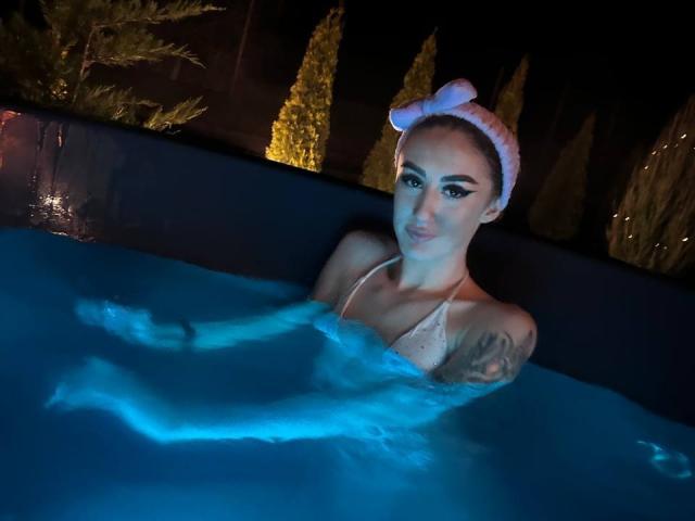 MackenzieHart in a pool, blue-lit water, white bikini, pink headband, tattoo on right arm, smiling, breasts partially submerged, legs crossed, night setting, visible eyeliner, dark background.
