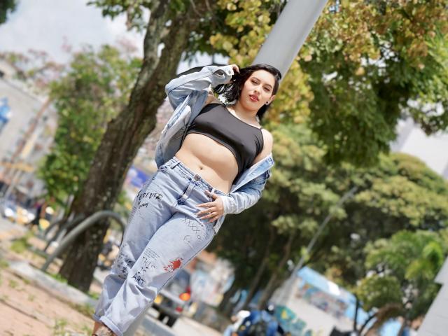 AmyRoss has dark hair, a black sports bra showing her tits, and light blue, ripped jeans with a jacket off her shoulders. She's standing in a park, looking sexy.