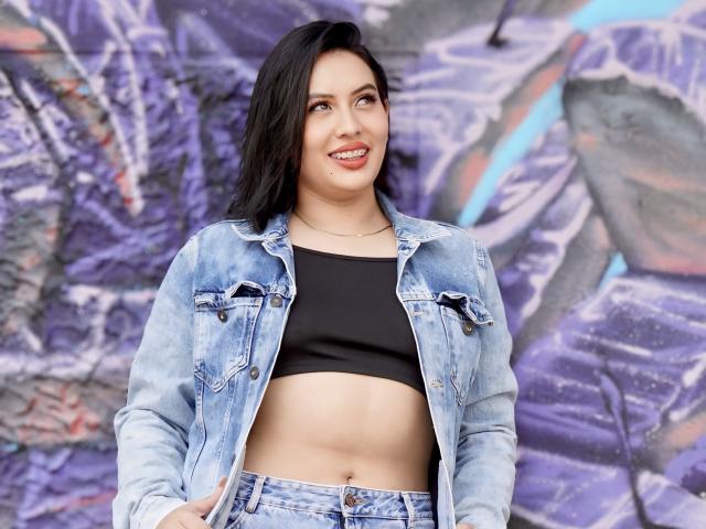 Amy Ross a les cheveux noirs, un sourire, une veste en jean et un crop top noir, qui dévoile sa poitrine et son nombril. Elle se tient devant une œuvre de graffiti..