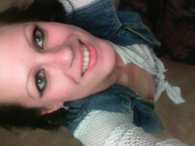 LussyKlara with a big smile, green eyes, and short dark hair, wearing a denim vest over a white knitted top, showing her small tits and nipples.