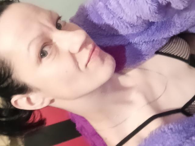 LussyKlara with fair skin, black bra, and mesh, wearing a purple fuzzy jacket, has short brown hair, and a neutral expression, showing her small tits and slim neck.