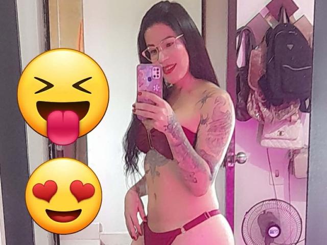 SexyCaandydoll with long black hair, glasses, red bra, and red panties, holding phone, tongue out emoji, heart emoji, tattoos on arm, standing in room with mirror, fan.