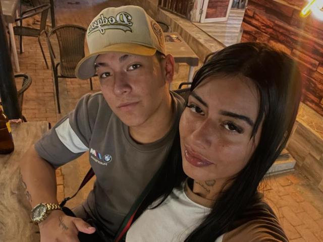 A selfie of ZairaAndres and a man in a dimly lit bar. ZairaAndres has long black hair, tan skin, and a nose piercing. She's wearing a white shirt. The man has a fair complexion, short brown hair, and wears a beige "Copa" cap, a grey shirt with a small logo, and a wristwatch. They're sitting close together at a wooden table. The man's left arm is tattooed. Warm, brown-toned background with brick walls and wooden textures. Both have neutral expressions. The man's cap is slightly worn. The image is clear with natural lighting.