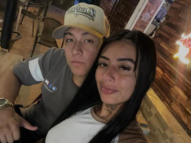 ZairaAndres with puffy ass and small tits, wearing a white shirt, next to a dude in a grey t-shirt and "Capo's" cap, in a dimly lit bar.