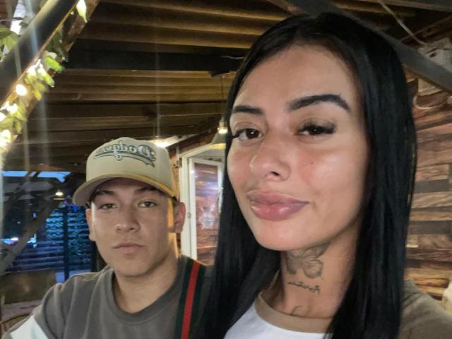 ZairaAndres, a young woman with long black hair, full eyebrows, and a tattoo on her throat, takes a selfie. She has light brown skin, subtle pink lipstick, and a slight smile. Behind her, a young man with light skin, brown eyes, and a beige "Brooks" cap looks to the side. He wears a gray shirt and a red strap. The wooden interior has string lights and a door. The image is a close-up selfie.