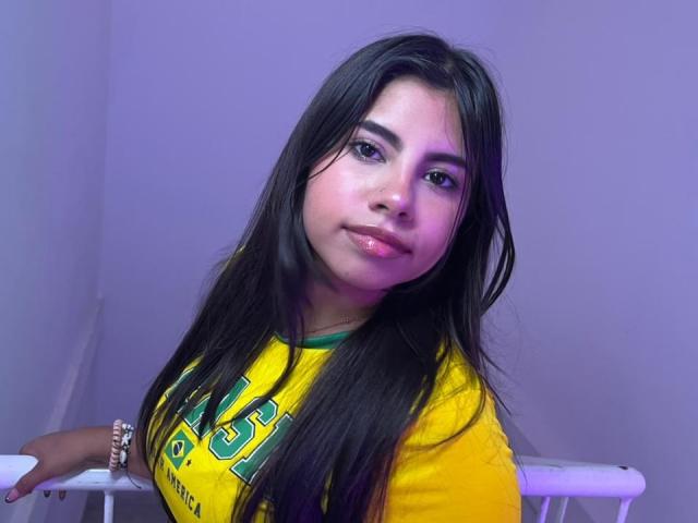 Celeste Morel, con lunghi capelli neri, labbra rosa lucide, indossa una maglia da calcio gialla, appoggiata a una superficie bianca, con una luce viola sul viso, che guarda verso la fotocamera..