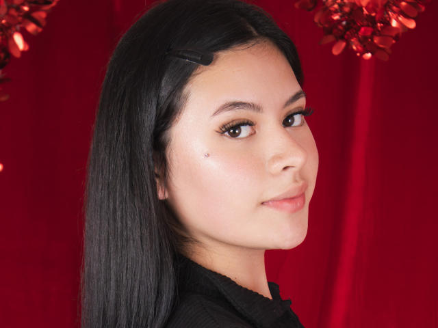 EimiLara with black hair, brown eyes, subtle makeup, small beauty mark on cheek, wearing black shirt, red background, slight smile, looking over shoulder, soft skin, cute face.