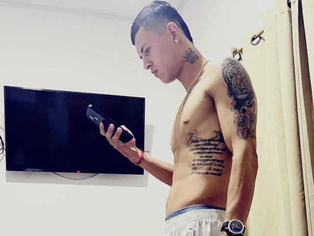 Shirtless TitanBodyX with tattoos, holding a game controller, looking down, wearing loose pants, standing in a room with a wall-mounted flat-screen TV.