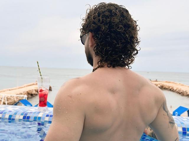 TatooMan, shirtless with curly brown hair, back to camera, standing in blue-tiled pool, water droplets on skin, tattoo on right arm, red drink with straw in foreground.