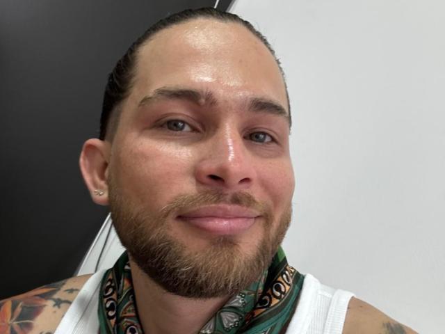 TattooMan has a shiny nose, brown beard, and tattoos on his arms. He's wearing a green patterned scarf, white sleeveless shirt, and has a small ear piercing.