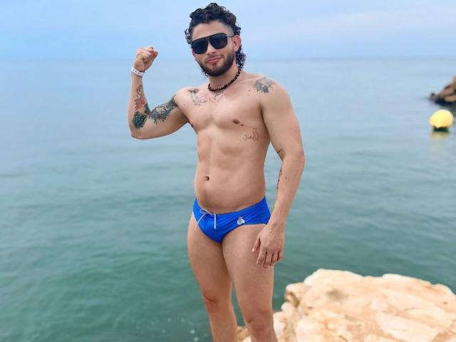 TattooMan, a muscular guy with tattoos, blue Speedo, and sunglasses, flexes his right arm on rocky beach, showing off his abs and a slight beard.