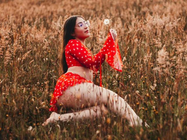 Ann Fisher, met lang zwart haar, een bril, een rode outfit met stippen, die een paardenbloem vasthoudt, knielend in een grasveld, waarbij haar ronde billen en blote benen zichtbaar zijn, in een sexy, natuurlijke pose..