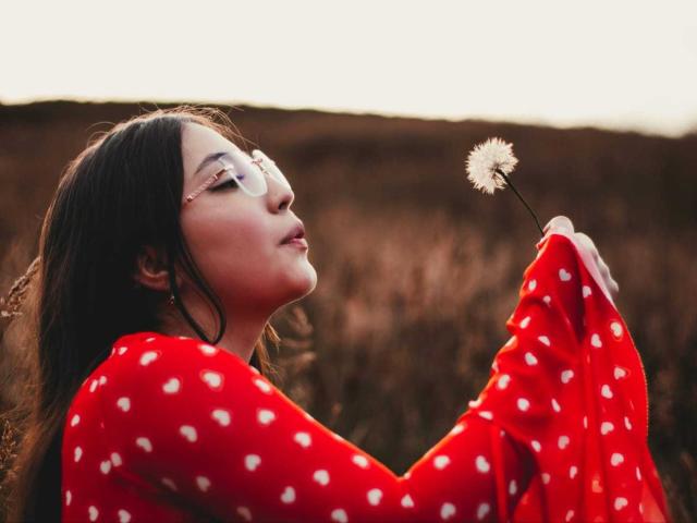 AnnFisher with long dark hair, glasses, and red polka dot sweater, holding a dandelion, eyes closed, smiling, in a sunlit field. Boobs not visible, focus on face.