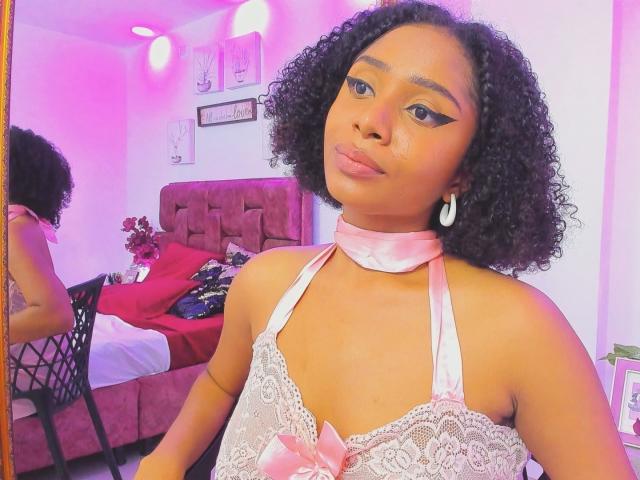 MeganWlliams with curly black hair, wearing a pink satin choker and lace dress, showing her small tits, in a pink-lit room, reflected in a mirror.