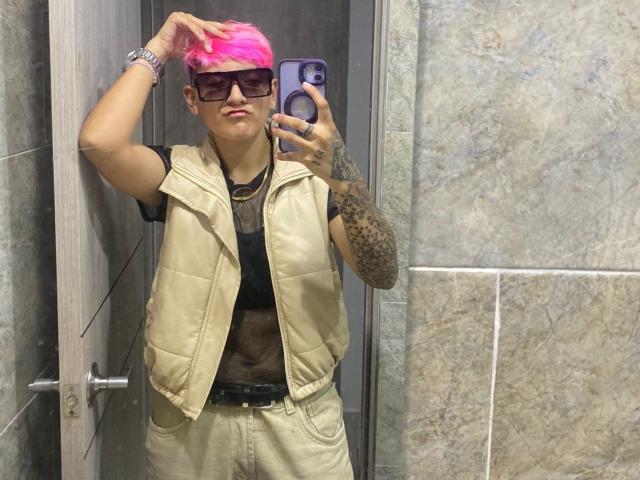 DannyRouds, con cabello rosa, gafas, tatuajes, chaleco beige, camisa negra, pantalones beige, tomando un selfie en un baño, con la mano en la cabeza, sonriendo, sosteniendo un teléfono morado..