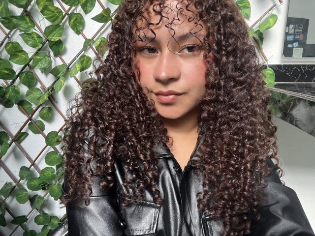 SofiaPassionne with curly brown hair wearing a black leather jacket, her face is slightly flushed, and she's looking directly at the camera with a subtle smile.