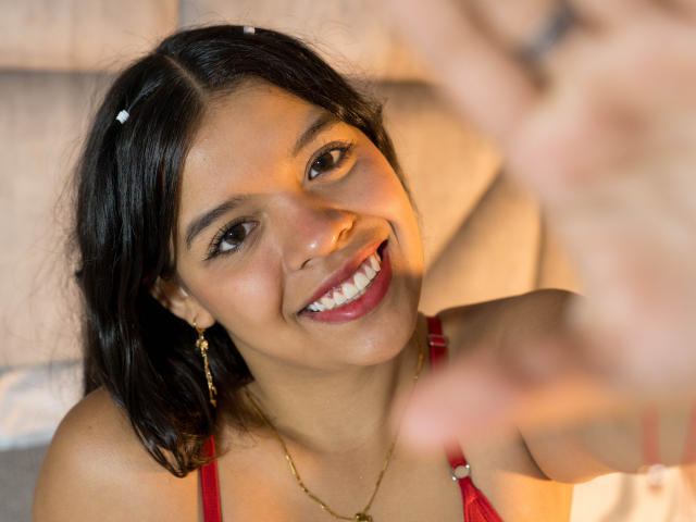 LiinaSantacruz has medium brown skin, dark hair, and is smiling with red lipstick. She's wearing a red top and gold earrings, with a blurred hand in the foreground.
