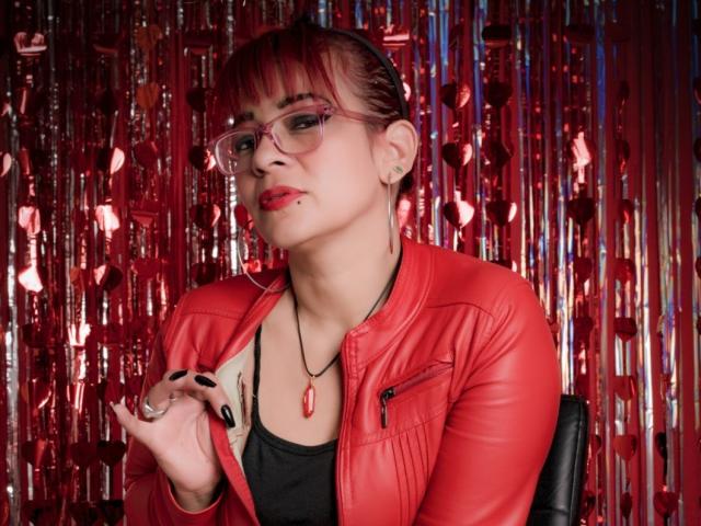 MissMirandaa met rood haar, een bril, een rode leren jas, een zwarte top en een rode ketting, met haar hand omhoog, waarbij ze zwarte nagels laat zien en een rode piercing in haar lip heeft..
