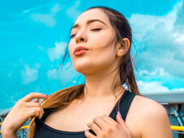 TaniaSandersX with closed eyes, brown hair in a ponytail, wearing a black tank top, pulling hair, under bright blue sky, looking confident, slightly pouty lips, visible nose.