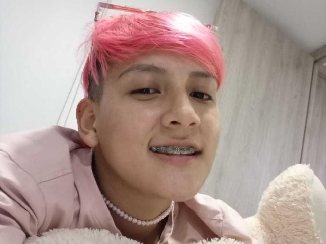 DannyRouds with pink hair, wearing a pink shirt and pearl necklace, smiling with metal teeth, holding a beige teddy bear.