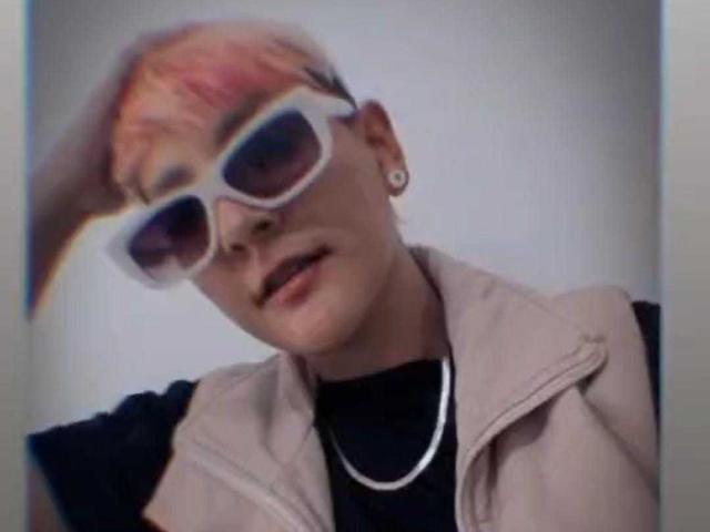 DannyRouds with pink hair, white sunglasses, and a beige vest, wearing a black shirt and silver chain, looking slightly to the side.