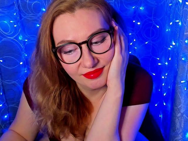 PaulinaSunny with red lipstick, black glasses, and wavy blonde hair, smiling, touching her cheek, blue-lit background, wearing a black top, showing her cute face and tits.