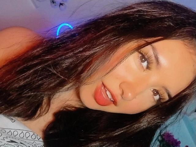 SusanMonroy has wavy dark brown hair, tan skin, and is sticking out her tongue with her pink lips slightly parted. Her brown eyes are looking sideways.