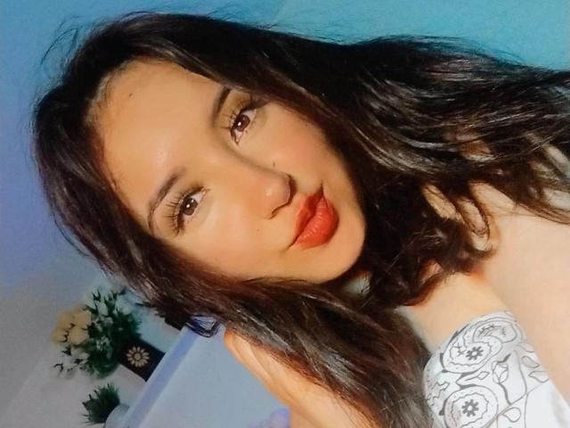 Susan Monroy, con lunghi capelli neri ondulati, occhi marroni e rossetto rosso, indossa una camicetta bianca con un motivo floreale, mostrando un viso carino e con le labbra leggermente schiusse, in una stanza ben illuminata..