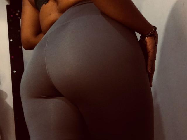 ShamanthaFire's big, round ass in tight, gray, butt-hugging leggings, with her hand on her hip, wearing a dark sports bra, dimly lit, showing her curvy, dark-skinned body.