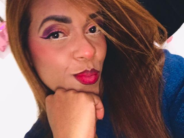 ShamanthaFire with her hand on her chin, sporting pink eyeshadow, purple lipstick, red lipstick, and brown hair, wearing a blue shirt, smiling slightly.