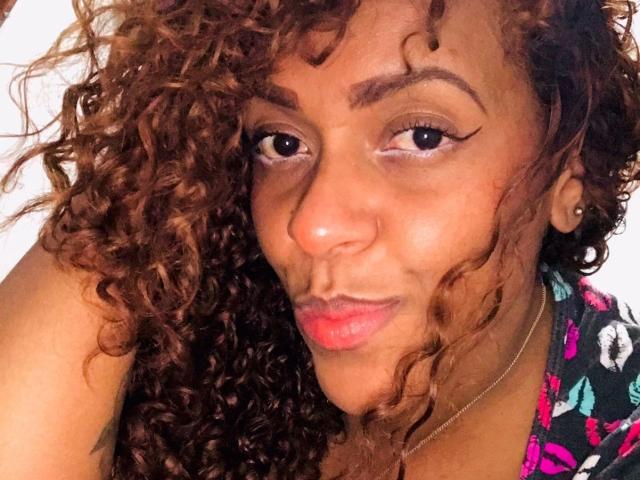 ShamanthaFire with curly brown hair, brown skin, and red lipstick, wearing a colorful patterned top, has a pouty face and winged eyeliner, looking slightly confused.
