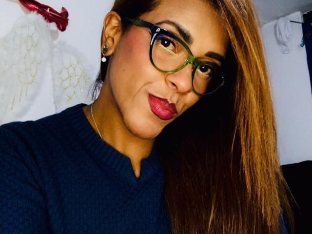 ShamanthaFire with green glasses, red lipstick, and dark blue sweater, showing off her tits and cute face in a selfie. Brown hair, ear piercings, and a slight smile.