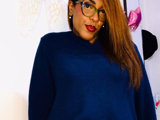 ShamanthaFire with long brown hair, green glasses, red lipstick, and a blue sweater, showing a slight smile, has a curvy ass, and is standing in a bright room.