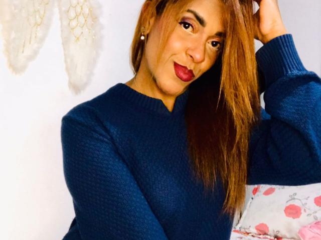 ShamanthaFire with long brown hair, dark blue knit sweater, red lipstick, smiling at camera, hand in hair, white wall, angel decor in background, sitting on pink floral pillow.