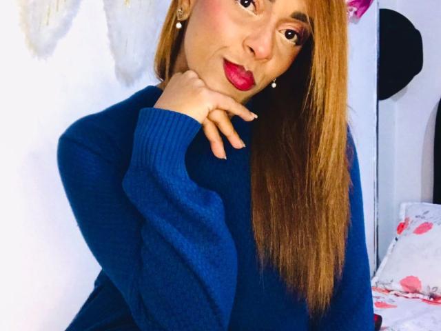 ShamanthaFire with long brown hair, wearing a blue sweater, red lipstick, and pearl earrings, touching her chin with a slightly pouty expression. Her tits are not visible.