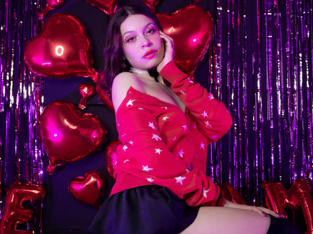 Sophie Blake, con occhi viola, un maglione nero con un motivo a stelle rosse, una gonna nera e uno sfondo viola lucido, appoggiata a palloncini a forma di cuore, con una mano sul viso, che guarda verso la fotocamera, illuminazione viola e rossa..
