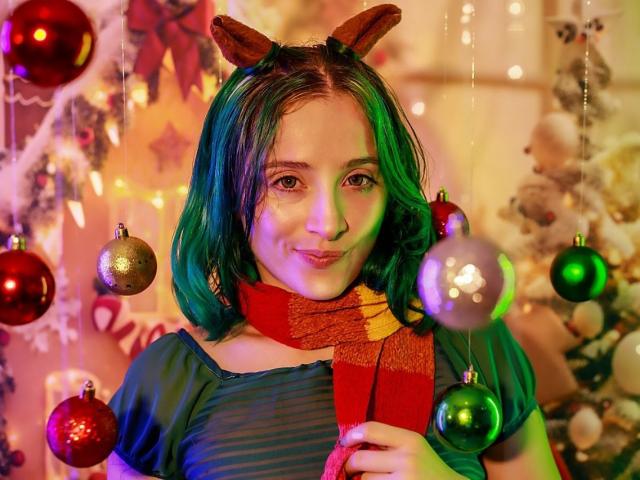 AshleySoy with green hair, wearing reindeer ears, red and yellow scarf, blue top, smiling, holding scarf, surrounded by colorful Christmas ornaments and soft lights.