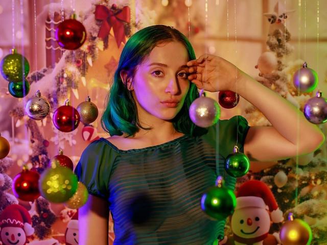 AshleySoy with green hair, wearing a sheer blue top, has one hand touching her forehead, surrounded by colorful Christmas ornaments and a decorated tree. Her tits are subtly visible.