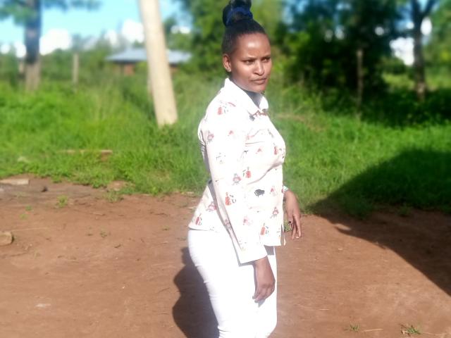 Emphi69 with dark skin, blue hair in a bun, wearing a white floral blouse and white pants, standing on a dirt path, looking over her shoulder, sunny day.
